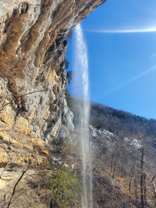 A waterfall cascades from a rocky cliff under a clear blue sky, surrounded by trees and mountains.